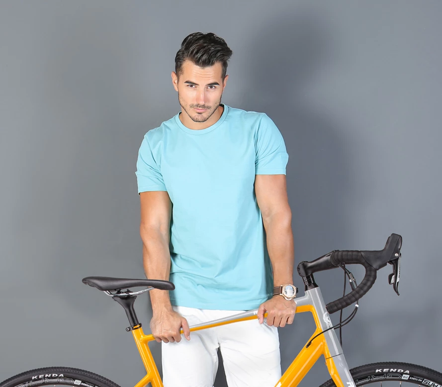 Man in light teal crew-neck t-shirt and white pants leaning on a yellow road bike against a gray studio backdrop