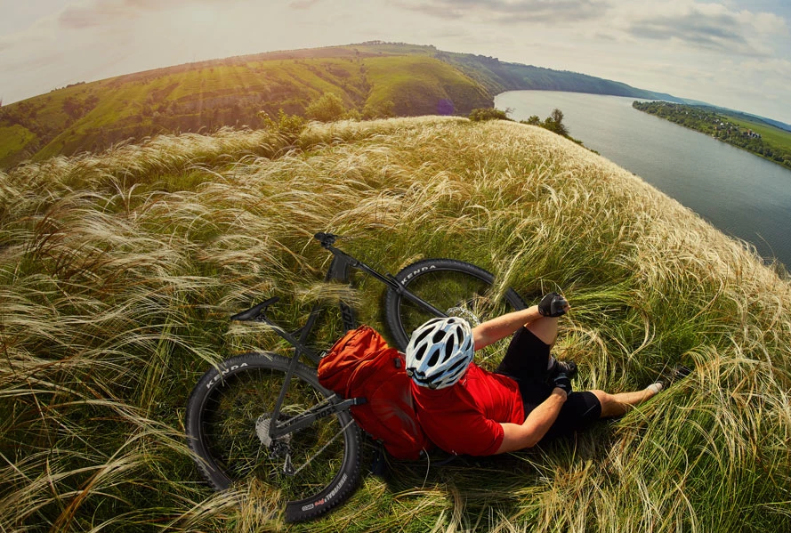 Mountain biker in red jersey and white helmet lying in tall grass on a hilltop beside a river, bike in foreground, fisheye