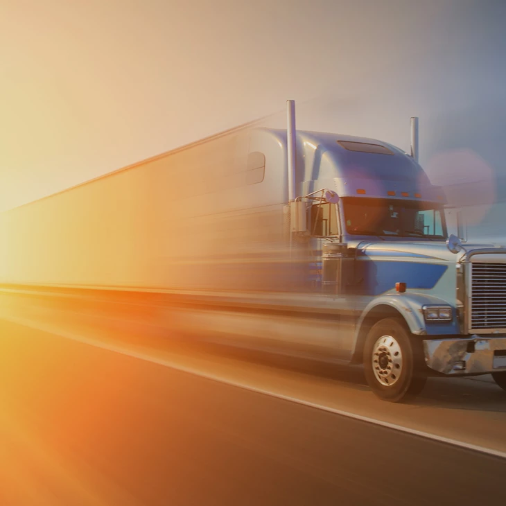 Blue semi truck with silver trailer speeding down highway at sunset, motion-blur effect with warm orange lens flare