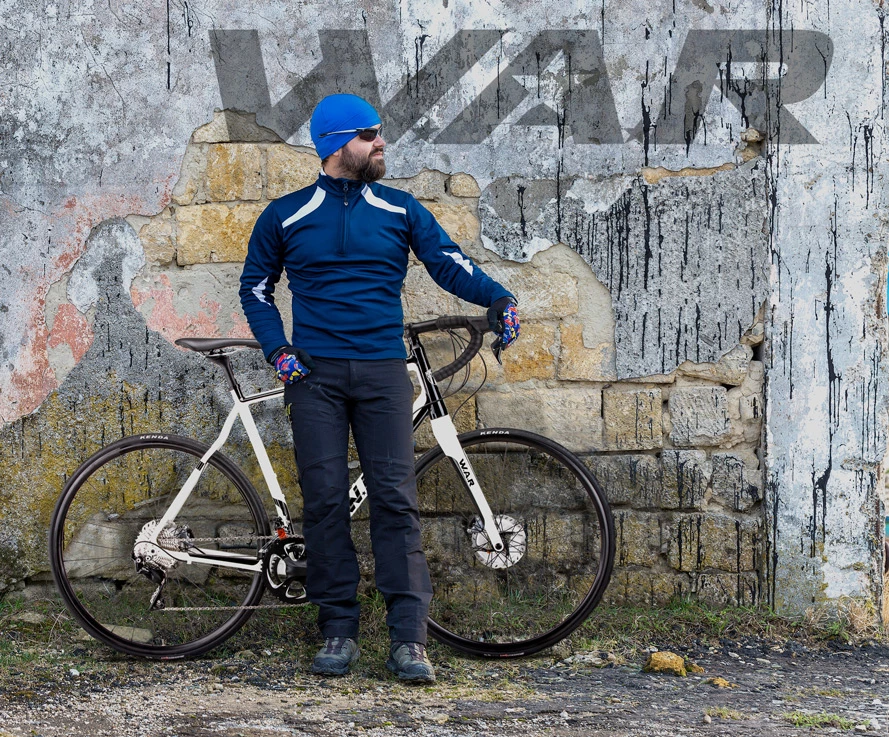 Bearded cyclist in blue beanie, sunglasses and navy zip-neck jersey standing with white road bike against weathered stone