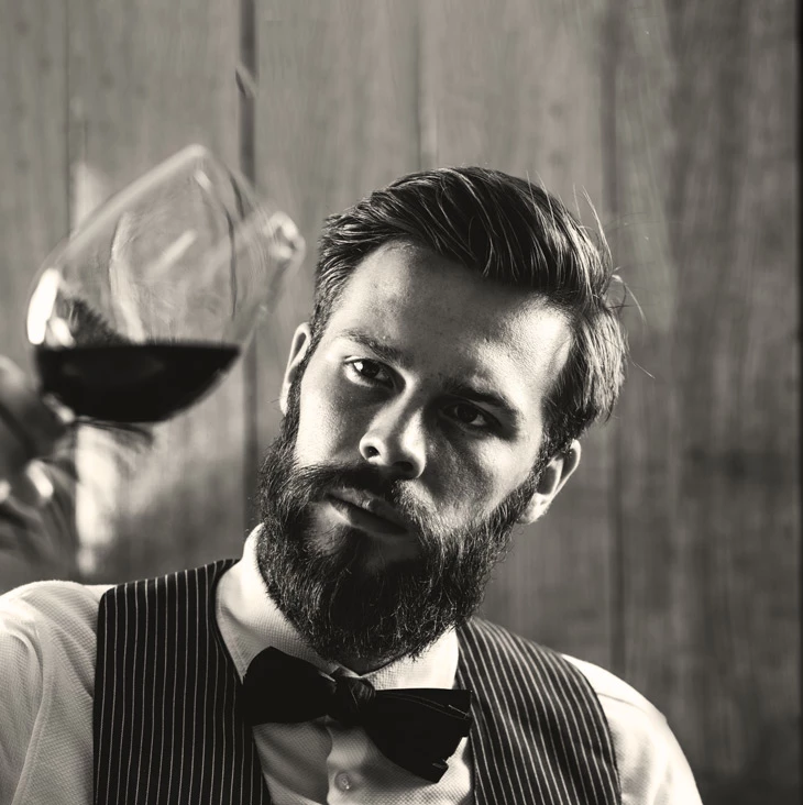 Bearded man in pinstripe vest and bow tie holding a glass of red wine